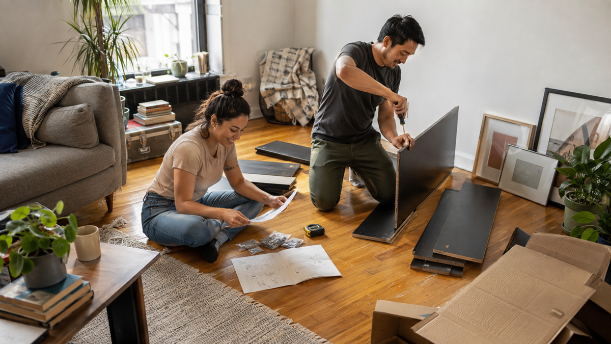 A couple assembles furniture on a wooden floor in a cozy living room, using instructions and tools. Panels, screws, plants, books, and framed art are scattered around them, creating a homey and collaborative atmosphere.