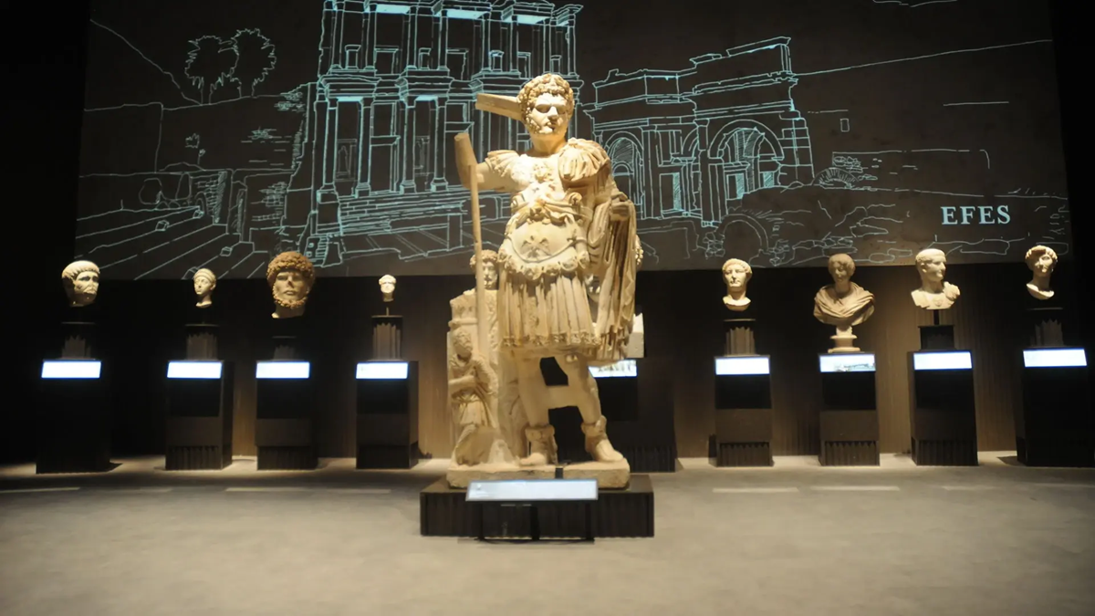 A large ancient statue stands center stage, surrounded by busts on pedestals. Behind them is a glowing outline drawing of ancient architecture labeled “EFES.” The setting appears to be a museum exhibit.