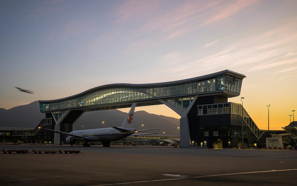 Airport at sunset with a modern, elevated glass terminal, a parked airplane on the ground, and another plane taking off in the sky. Mountains are visible in the background.