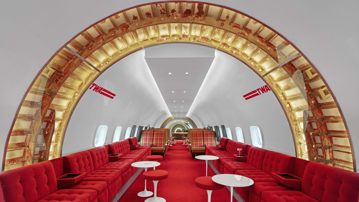 Retro airplane lounge with red seating, white tables, gold arched ceiling, and TWA logos on the walls. The design features a vintage, luxurious atmosphere inside the aircraft.