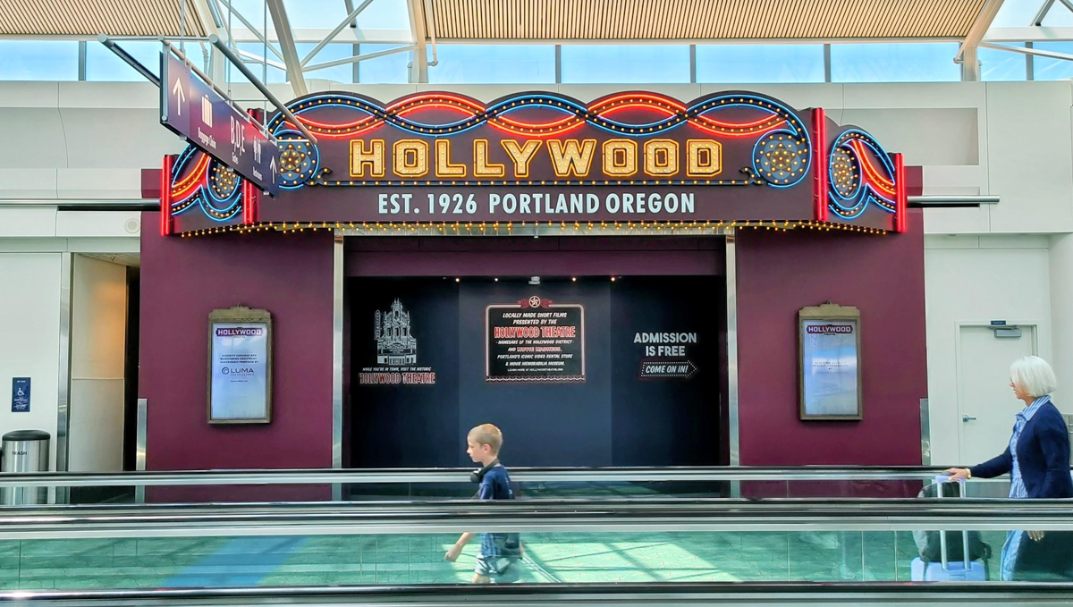 A boy and a woman walk past a vintage-style “Hollywood” sign display reading “Est. 1926 Portland Oregon” inside an airport with bright lighting and moving walkways.