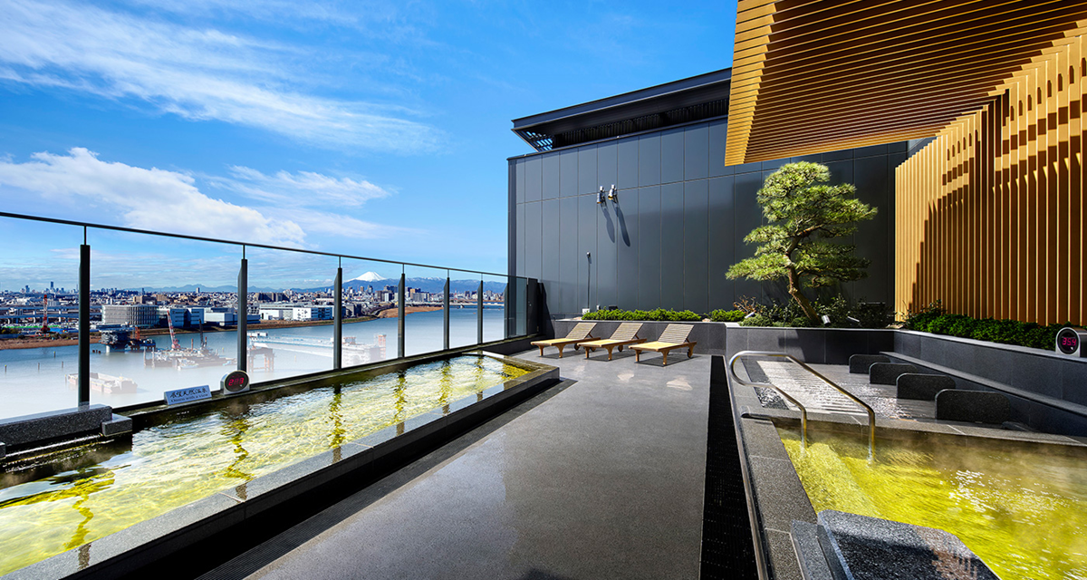 A modern outdoor hot spring bath with yellow-tinted water, lounge chairs, glass railings, city views, and a small tree under a blue sky.