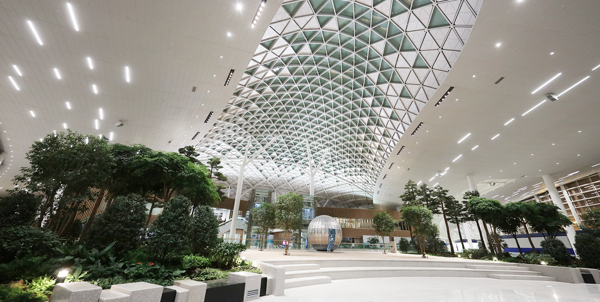 Spacious, modern airport terminal with a high, curved glass ceiling, lush indoor trees and plants, white floors, and ample lighting, creating an airy and welcoming atmosphere.