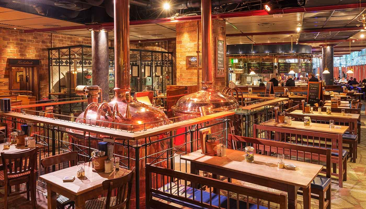 A spacious restaurant with wooden tables and chairs, copper brewing tanks in the center, and brick walls. The atmosphere is warm and inviting, with soft lighting and a mix of open and cozy seating areas.