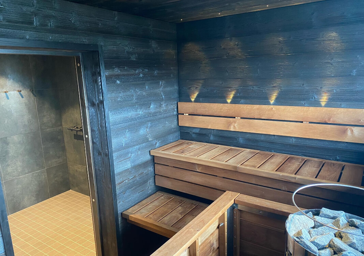 Wooden sauna interior with benches, a bucket of rocks, and dark wood-paneled walls. An open door leads to a tiled shower area with hooks on the wall. Warm lights illuminate the sauna walls.
