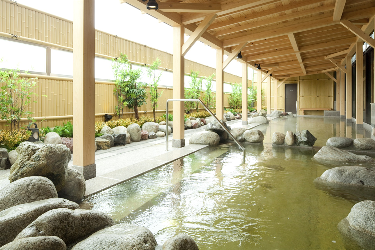 A serene indoor-outdoor Japanese onsen bath with clear water, natural rocks, bamboo plants, and a wooden roof structure, creating a peaceful and relaxing atmosphere.