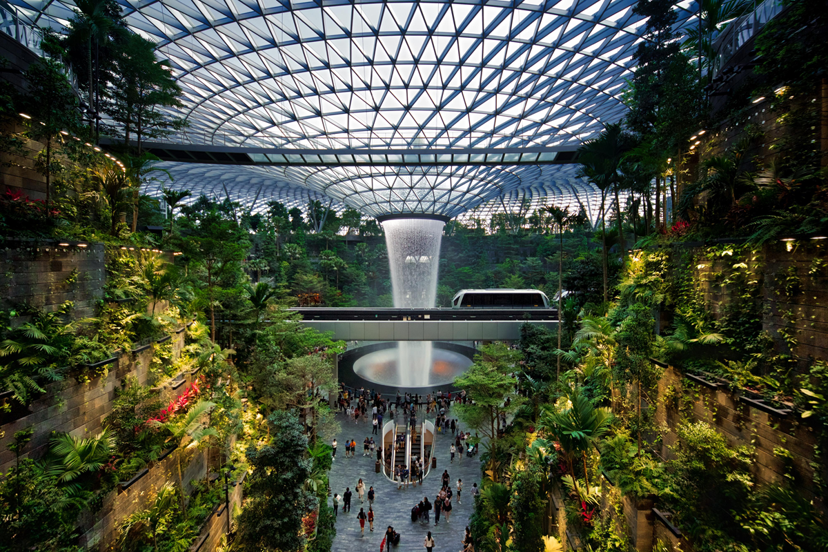 Indoor garden with lush greenery, a glass-domed roof, and a large central indoor waterfall surrounded by people on the ground level.