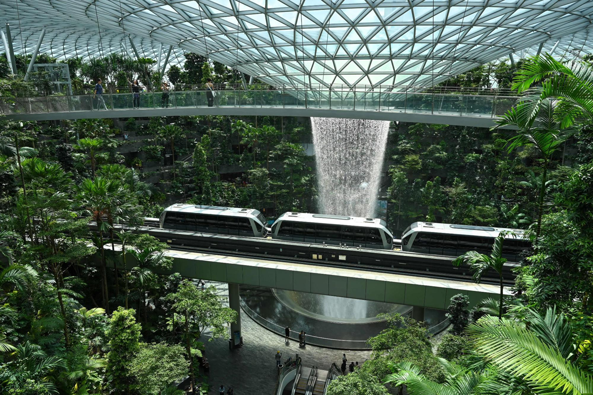 A train travels across a sky bridge in a lush indoor garden with a large waterfall and glass dome ceiling, surrounded by greenery and visitors walking below.