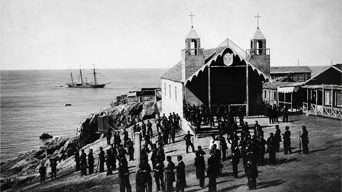 Historic black-and-white photo of soldiers gathered outside a church by the sea, with a sailing ship anchored offshore and wooden buildings nearby.