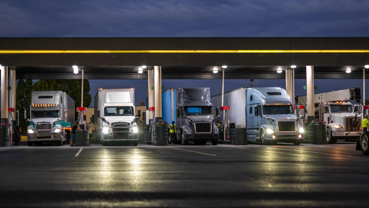 Six semi-trucks parked side by side at a truck stop fuel station at dusk, with headlights on and the sky darkening in the background.