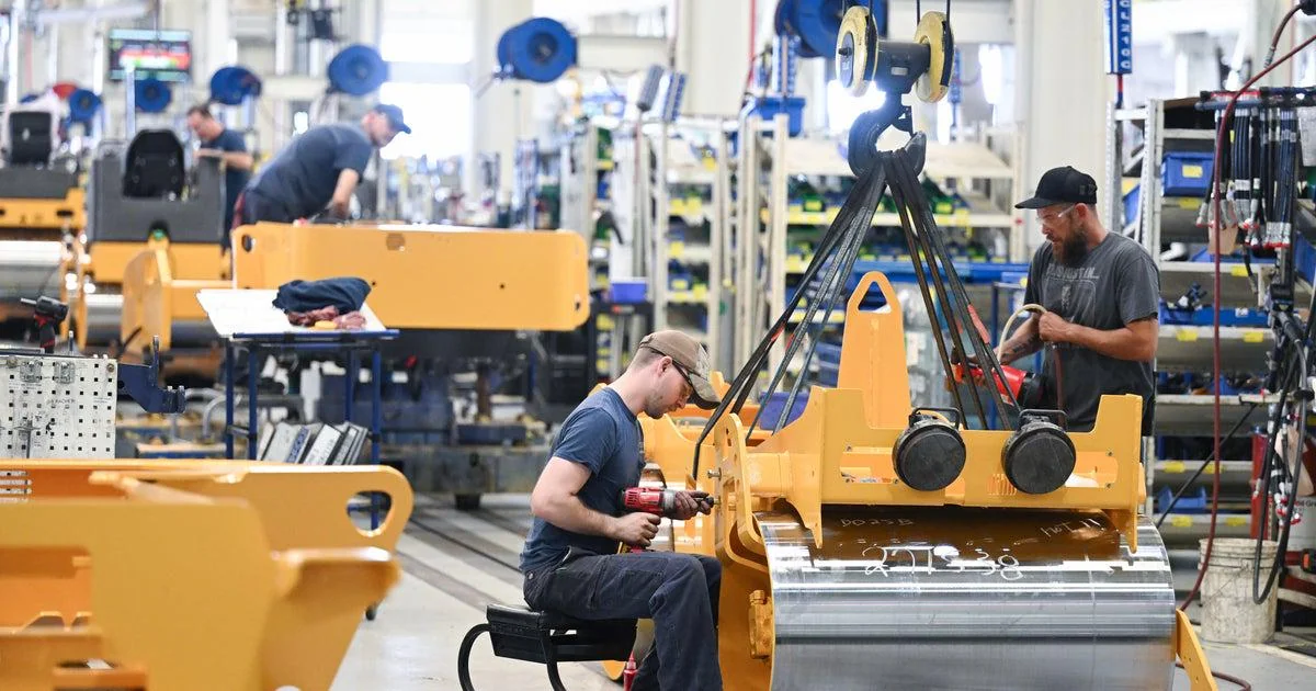 Workers assemble and work on large yellow machinery parts in a busy factory, using tools and wearing protective hats. Shelves and equipment are visible in the background.