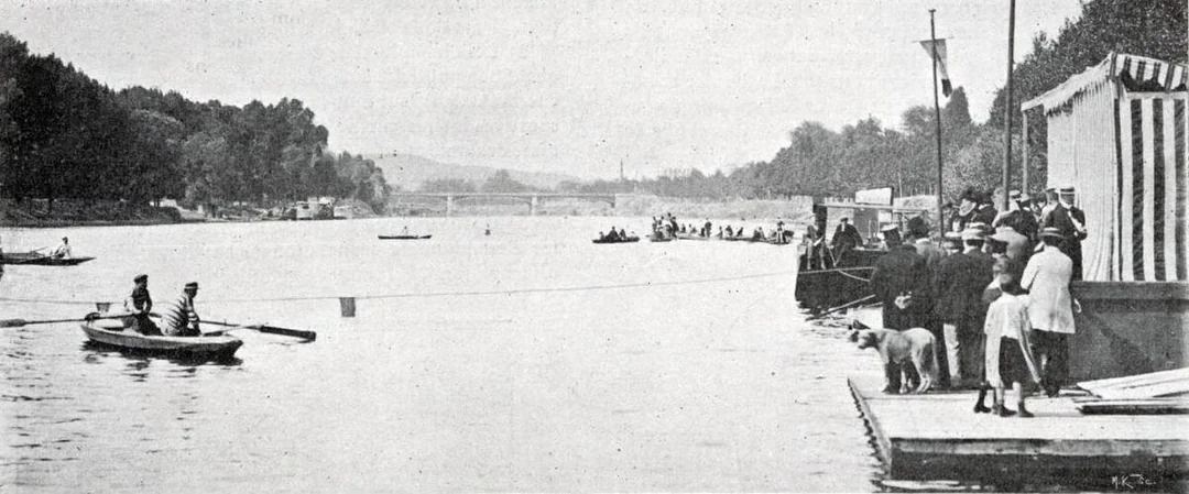 Black and white photo of people in early 1900s attire standing on a dock by a river. Rowboats and spectators are visible on the water, along with a striped tent and a dog on the dock. Trees line both riverbanks.