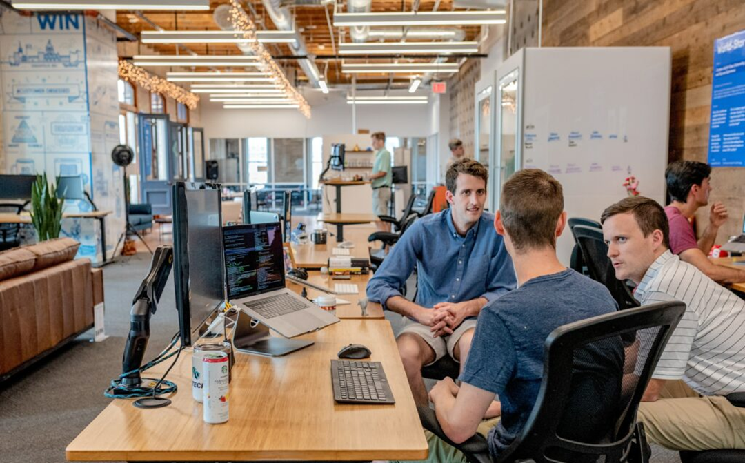 Three people sit at a desk with computers, engaged in conversation in a modern, open office. Other employees work in the background among bright lighting, whiteboards, and wooden decor.