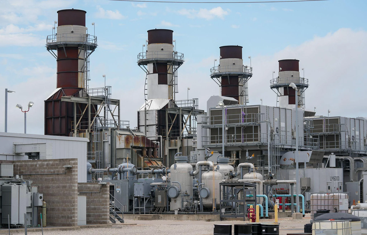 Industrial facility with multiple large smokestacks, pipes, and machinery. The sky is partly cloudy, and there are various metal structures and tanks throughout the site. No people are visible.