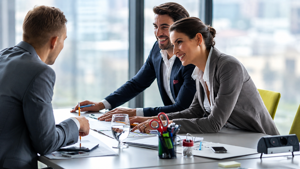 Three professionals in business attire sit at a desk in a modern office, smiling and engaged in conversation. Papers, pens, and office supplies are on the table, with large windows showing a city view in the background.