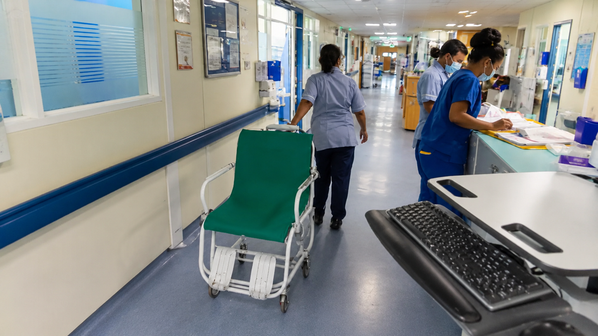 Hospital corridor with nurses in uniform working at a nurses’ station, a wheelchair in the hallway, and medical equipment nearby. Bright lighting and health-related posters are visible on the walls.