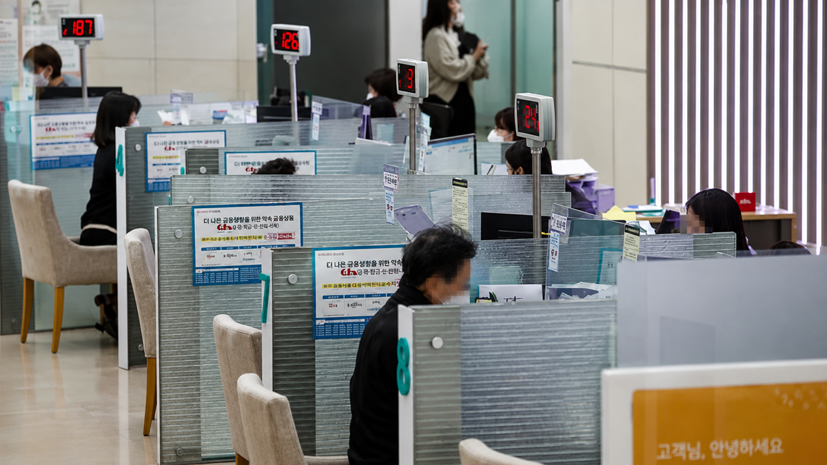 People sit at partitioned desks in an office, each with numbered signs. Some workers assist customers. Most people wear masks, and posters with information are attached to the partitions. Vertical blinds cover the windows.