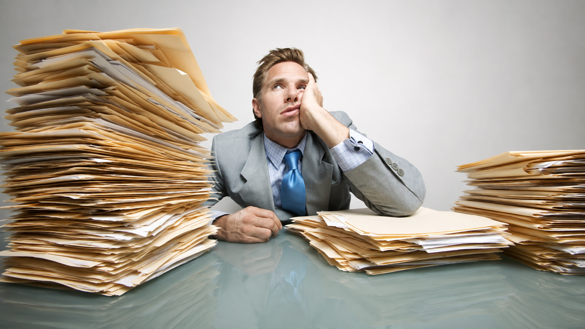 A man in a suit looks overwhelmed as he sits at a desk, resting his head on his hand, surrounded by large stacks of paperwork.