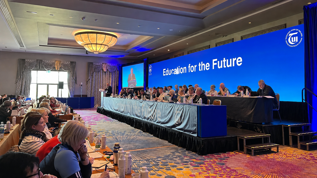Conference room with people seated at long tables facing a stage. A large blue screen reads “Education for the Future.” Several speakers sit at a head table, and attendees are watching and taking notes.