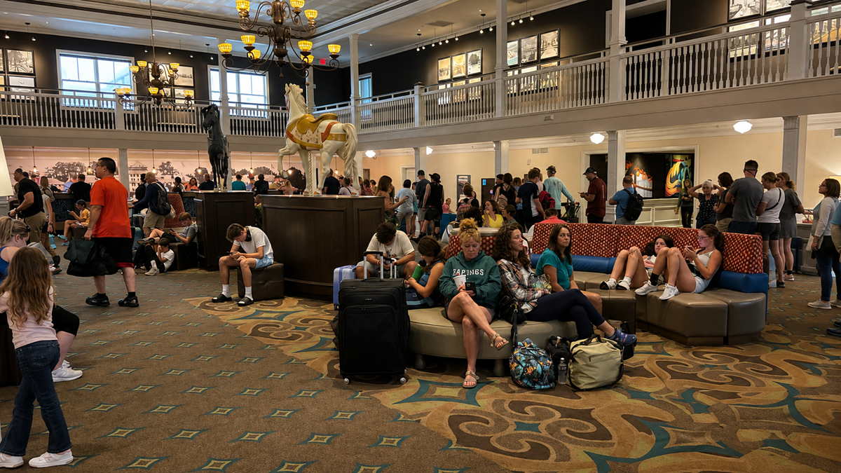 Large hotel lobby with many people sitting on couches and standing. A large white carousel horse is in the center. Overhead, ornate lights hang from the high ceiling. The atmosphere is busy and casual, with families and groups waiting.