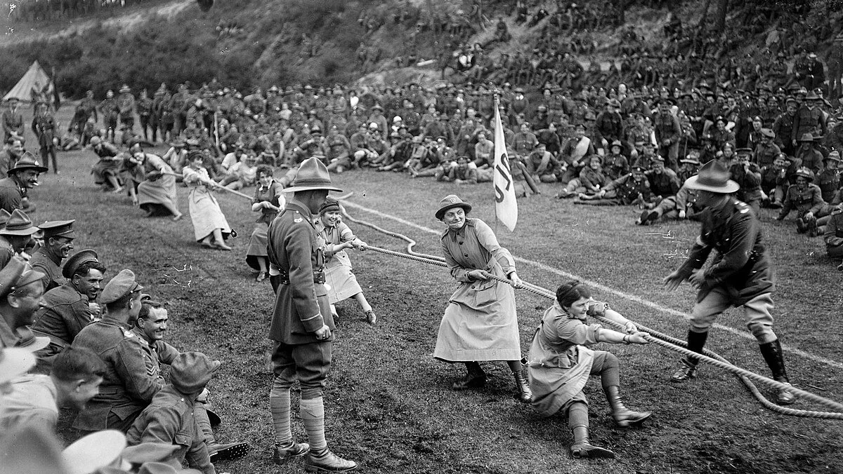 Women and men in early 1900s attire compete in a tug-of-war, with a large crowd of soldiers and spectators watching on a grassy field. sport