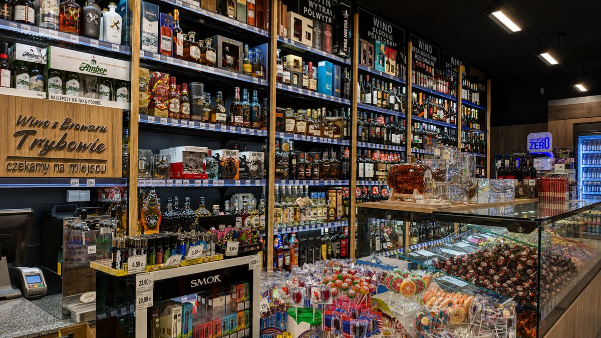 A liquor store interior with shelves filled with various bottles of alcohol, a display case of sweets, and colorful lollipops in the foreground. Signs and price tags are visible throughout the shop. Industry