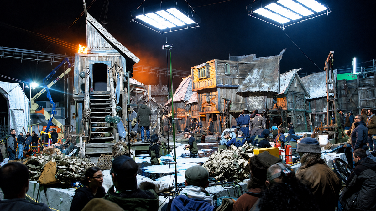 A busy movie set at night depicts a medieval village with wooden buildings, actors in costume, crew members, lighting rigs, and filming equipment visible throughout the scene.