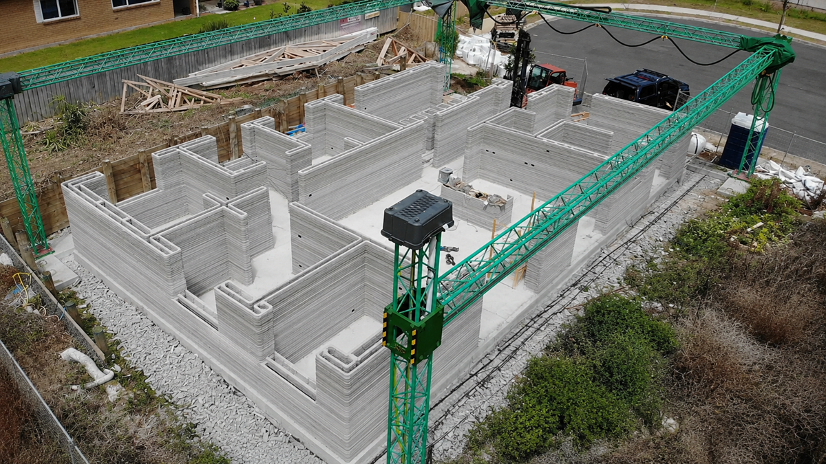 A green 3D printer constructs the concrete walls of a house on a building site, surrounded by grass, debris, and some vehicles parked on the nearby street.