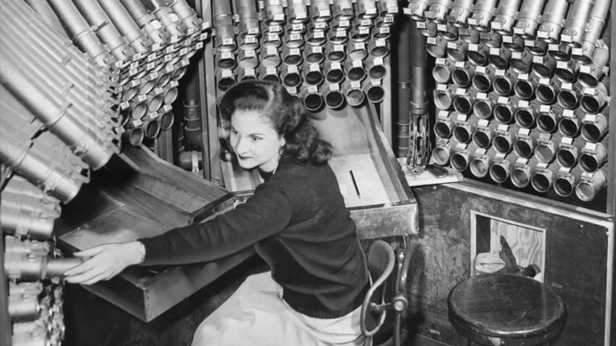 A woman sits at a control desk surrounded by numerous tubes or pipes, adjusting controls. The environment looks mechanical and complex, possibly related to an old machine or communication system.