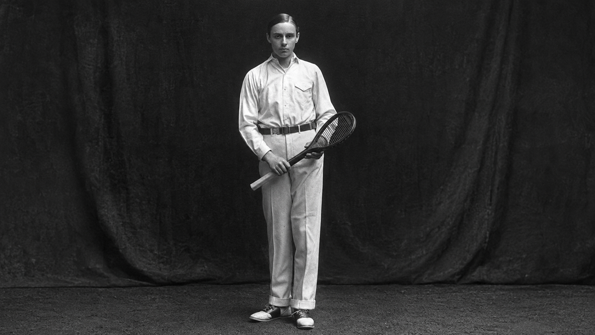 A person wearing light-colored, vintage tennis attire stands holding a tennis racket against a dark backdrop, looking directly at the camera.