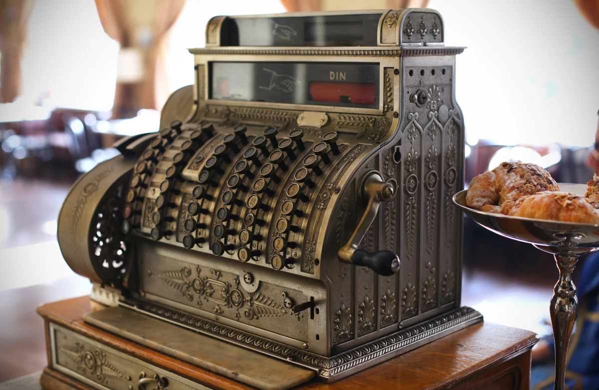 A vintage ornate cash register with detailed metalwork sits on a wooden counter next to a tray of pastries in a warmly lit room.