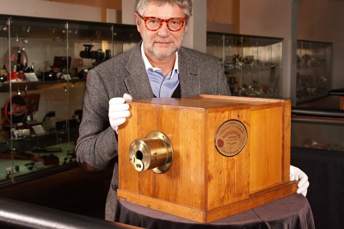 A man in glasses and a gray suit holds a large, antique wooden camera with a brass lens, displayed in a museum setting with glass cases in the background.