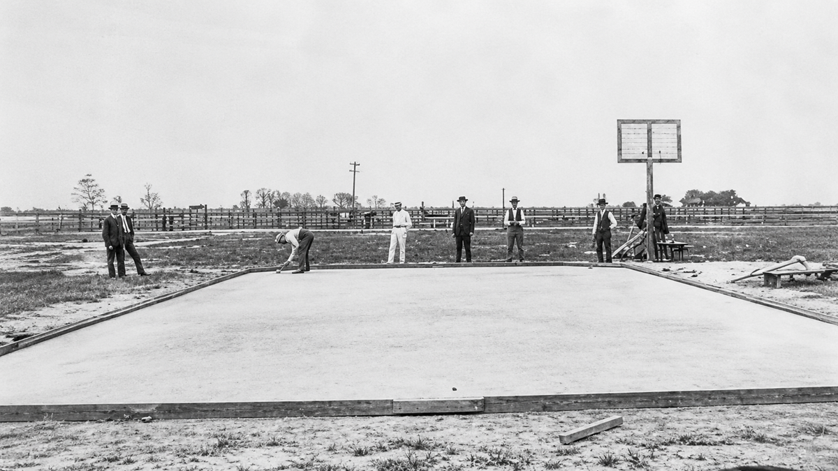 Seven men stand around a simple outdoor basketball court in a rural area. One is bending down, while others stand in the background. The court is unpaved with a single wooden backboard and hoop.