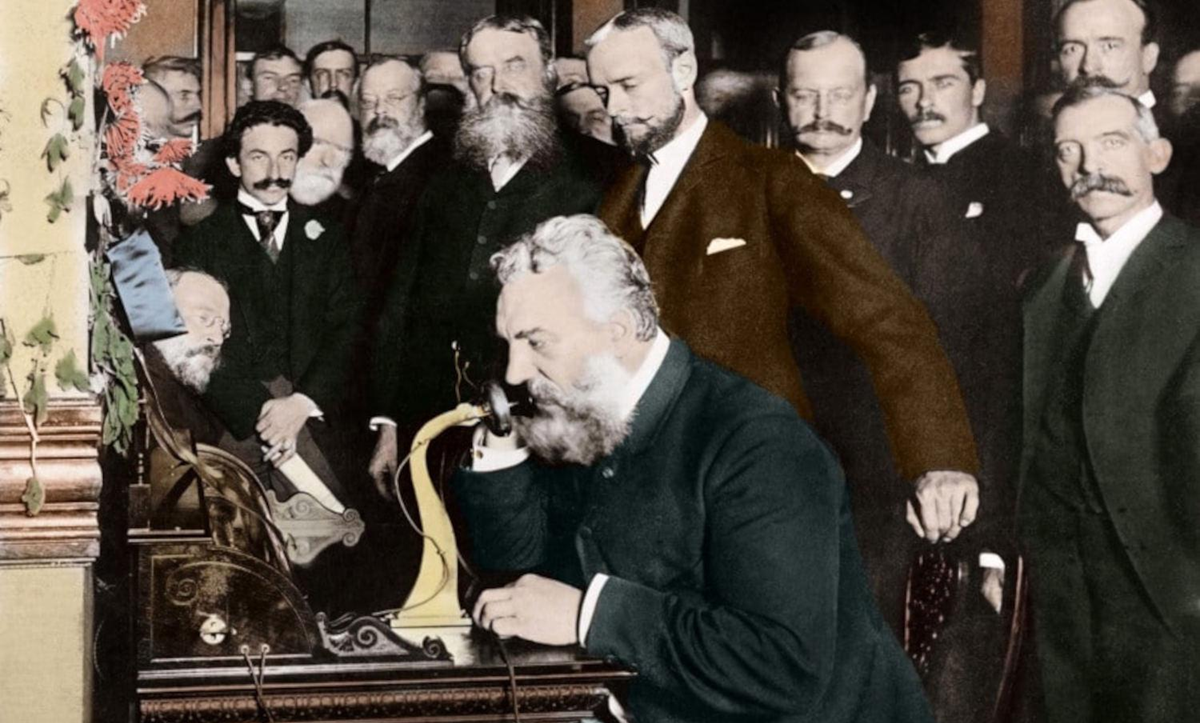 A group of men in old-fashioned suits watches as a man with a white beard speaks into an early telephone device in a historical setting.