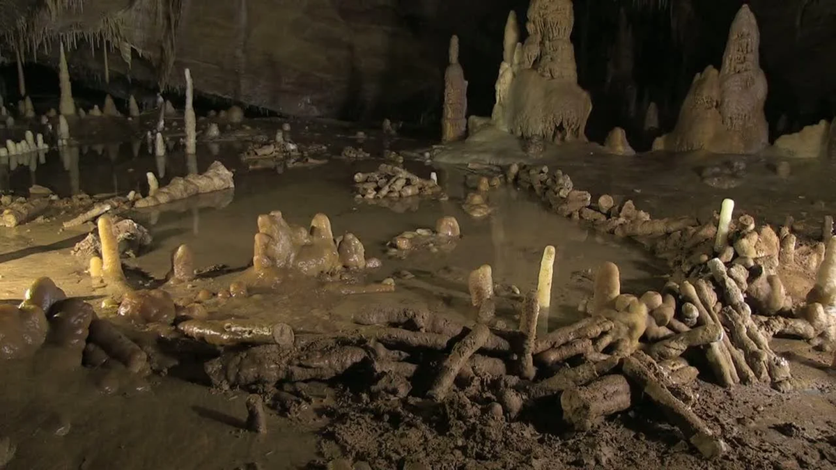 A cave interior with circular and semi-circular stone structures surrounded by stalagmites and stalactites, with muddy ground and reflective water patches.