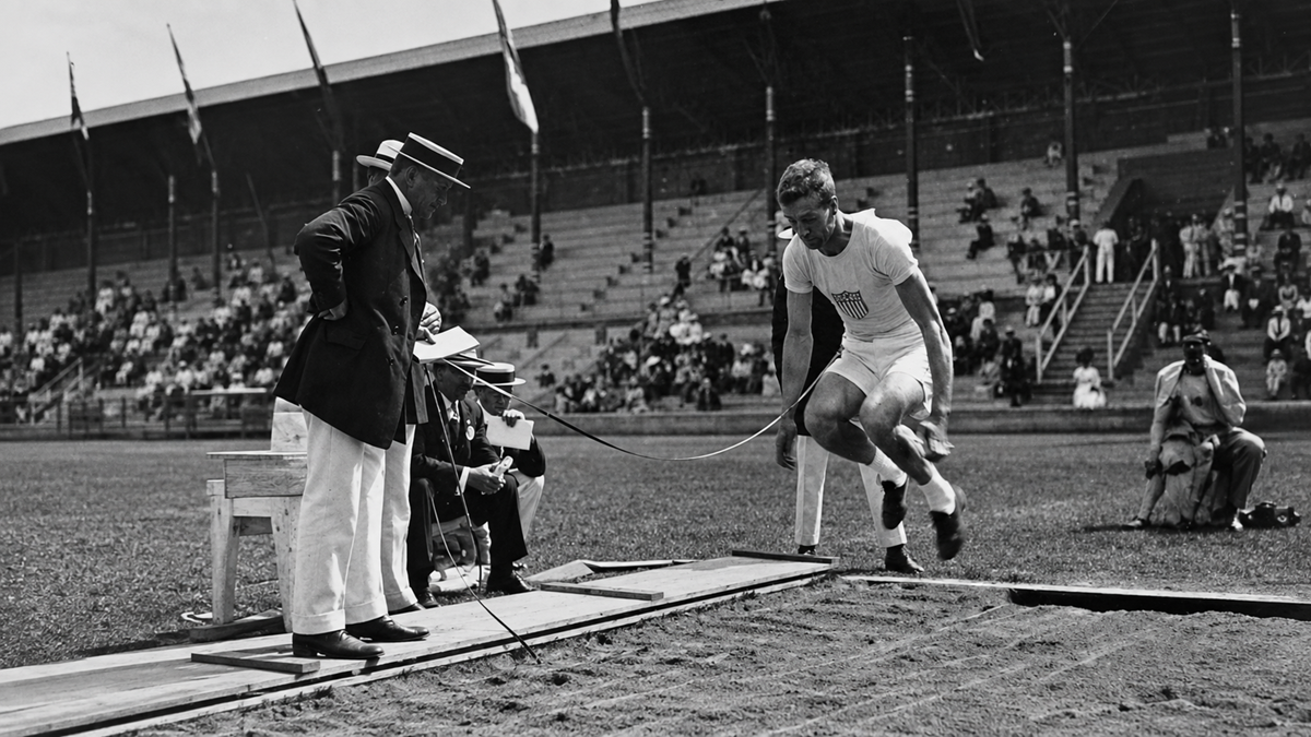 An athlete in mid-air attempts a long jump while officials observe closely at a stadium, with spectators seated in the background.