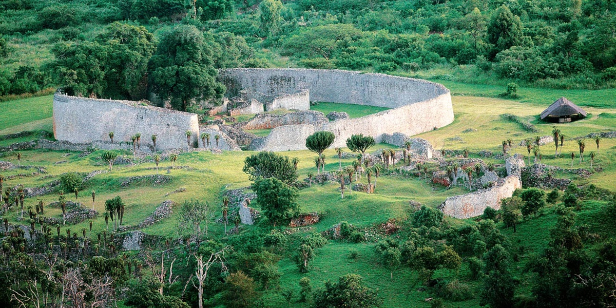 Ancient stone ruins surrounded by green vegetation, with large curved walls and scattered trees. A small thatched-roof hut stands to the right of the ruins, all set in a lush, hilly landscape.