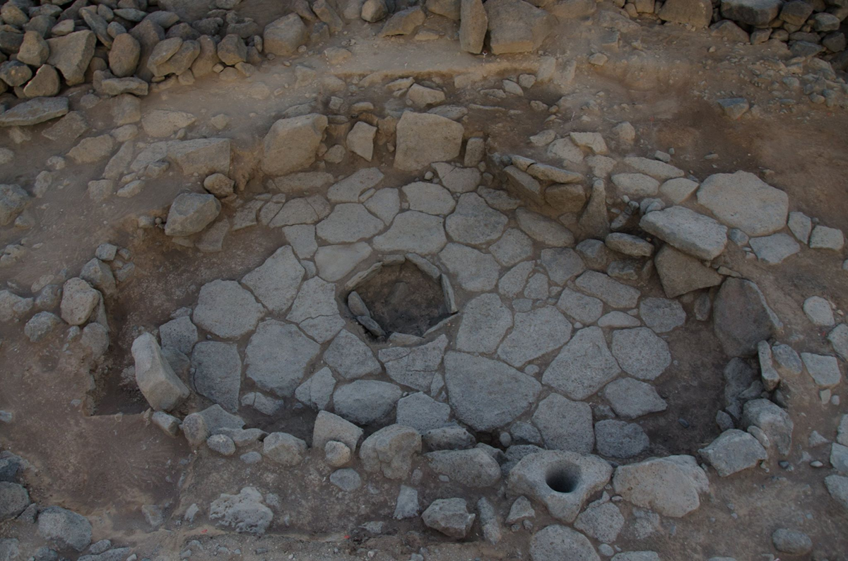 Ancient stone structure with a circular pattern, central hole, and surrounding rocks, possibly part of an archaeological site. Dirt and stones are visible around the patterned area.