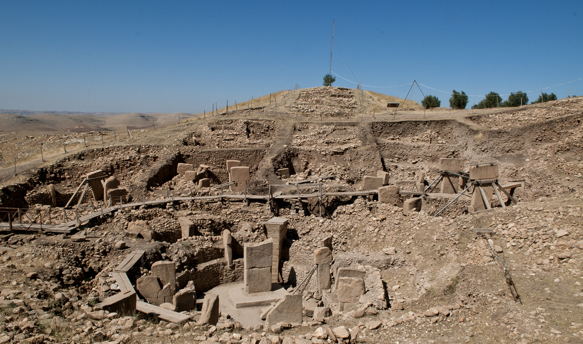 Archaeology. Ancient stone pillars and excavated circular structures at Göbekli Tepe archaeological site, surrounded by arid, rocky terrain under a clear blue sky. Wooden walkways cross the excavation area.