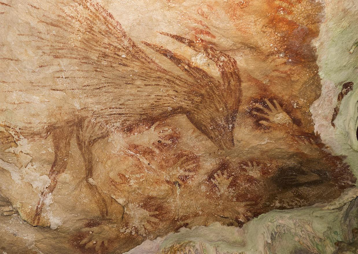 Cave painting showing a brown animal, possibly a goat or deer, surrounded by numerous hand stencils in red and brown tones on a textured rock surface.