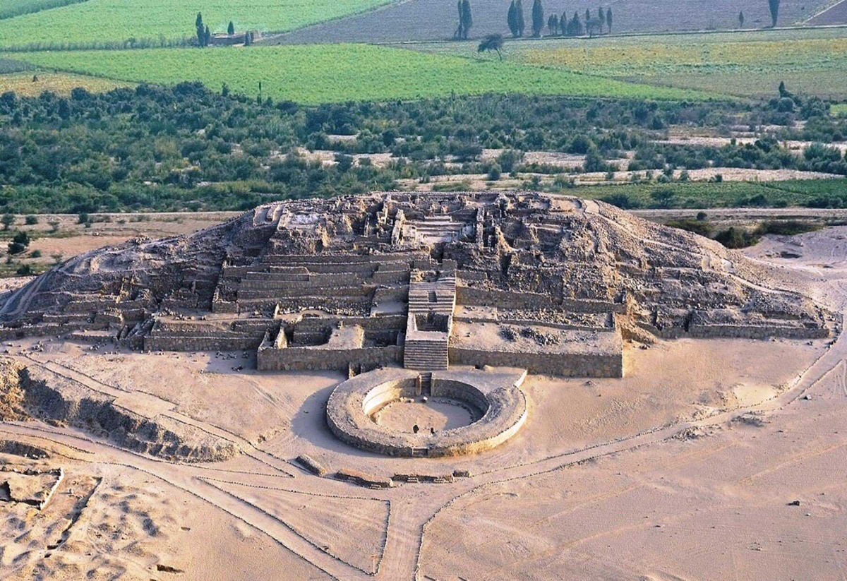 Aerial view of ancient stone ruins with a large circular structure in front, surrounded by desert sand, with green fields and trees in the background.