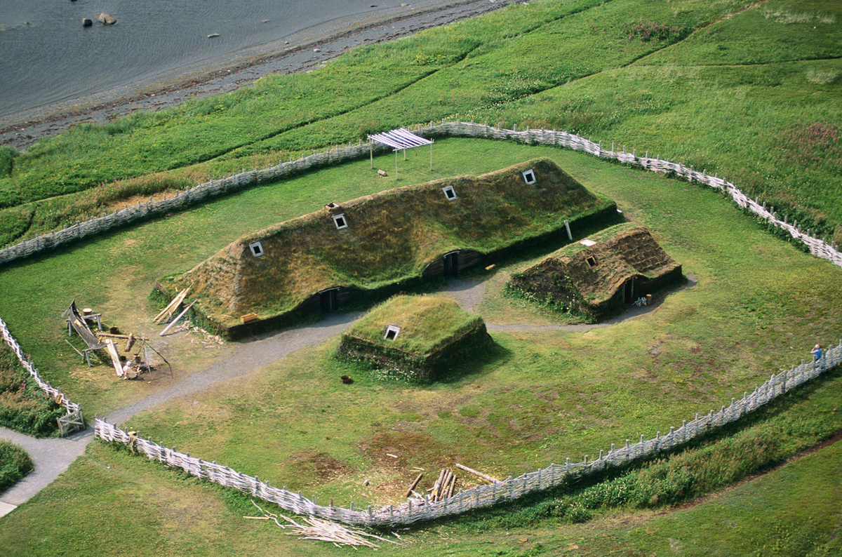 Aerial view of grass-covered Viking-style sod houses surrounded by a wooden fence, with paths, scattered wood, and greenery near a body of water.