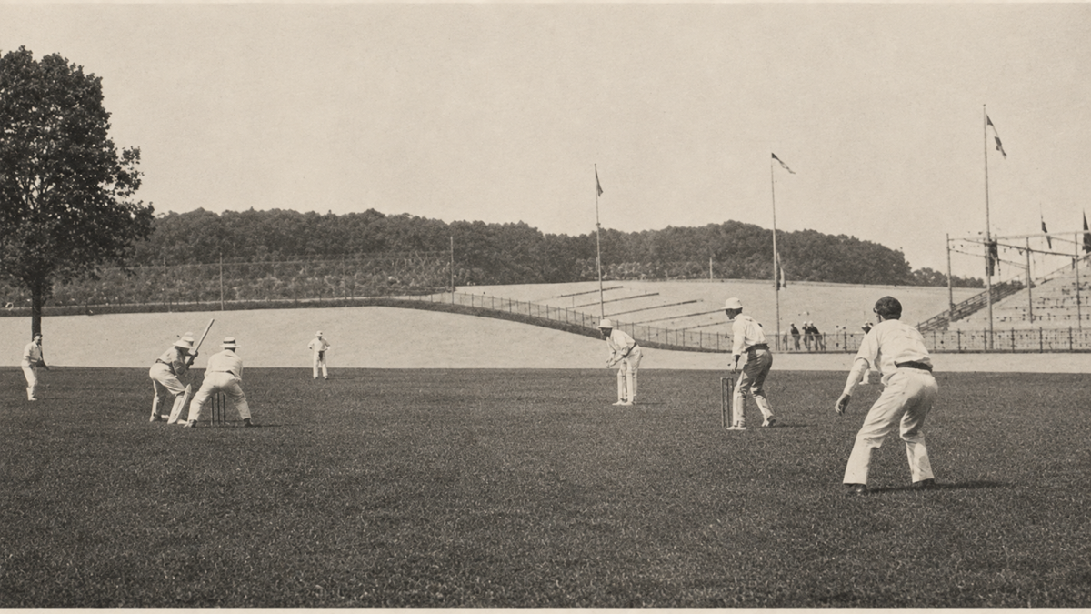 A vintage photograph of a cricket match being played on a grassy field with several players in white uniforms; empty bleachers and flags are seen in the background.