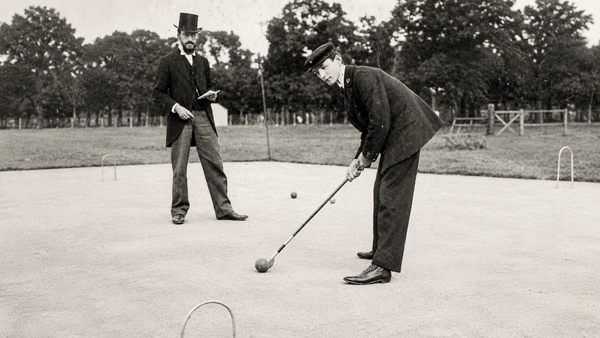 Two men in vintage suits and hats play croquet on a lawn. One man prepares to hit a ball with a mallet, while the other observes, holding a notebook. Trees and a fence line the background.
