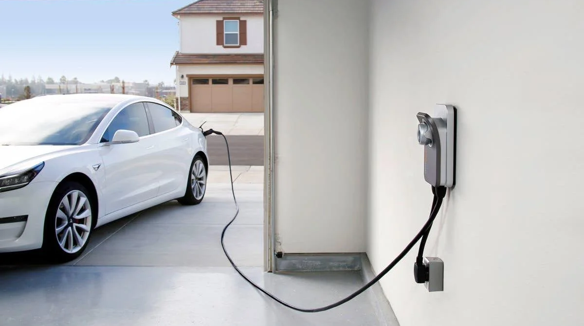 A white electric car is parked in a driveway, plugged into a wall-mounted home charging station inside a garage. A suburban house and clear sky are visible in the background.