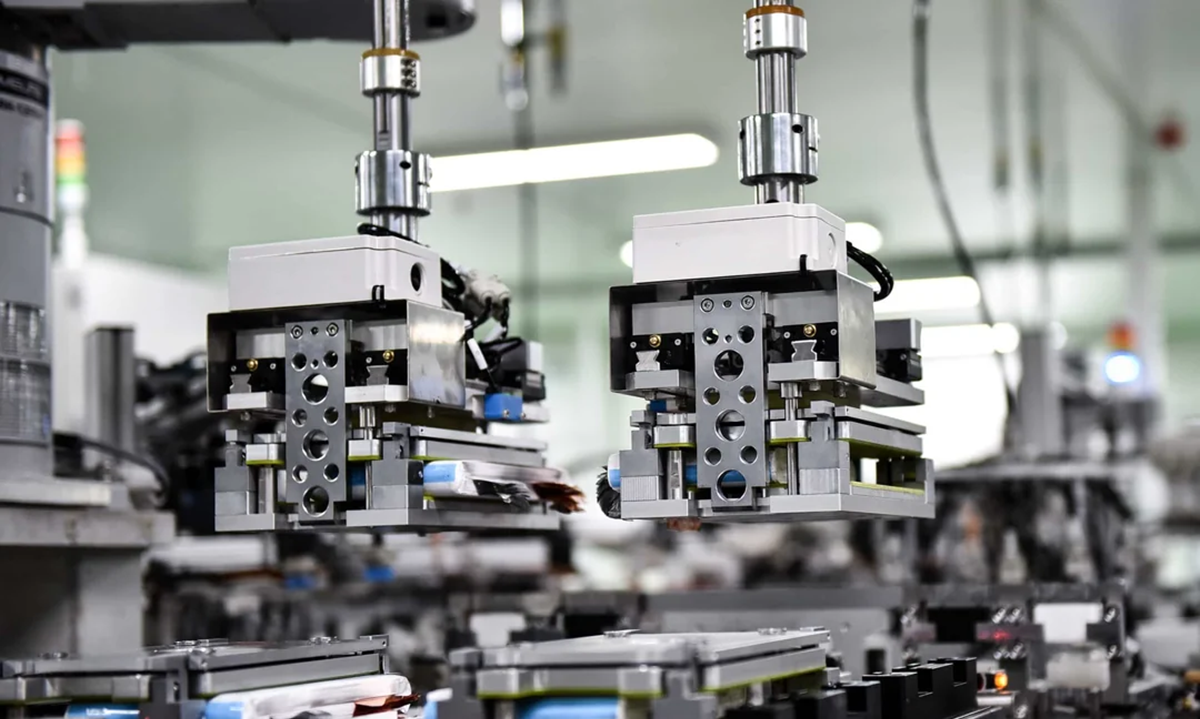 Close-up of automated robotic arms assembling electronic components in a factory setting, with blurred machinery and equipment visible in the background.