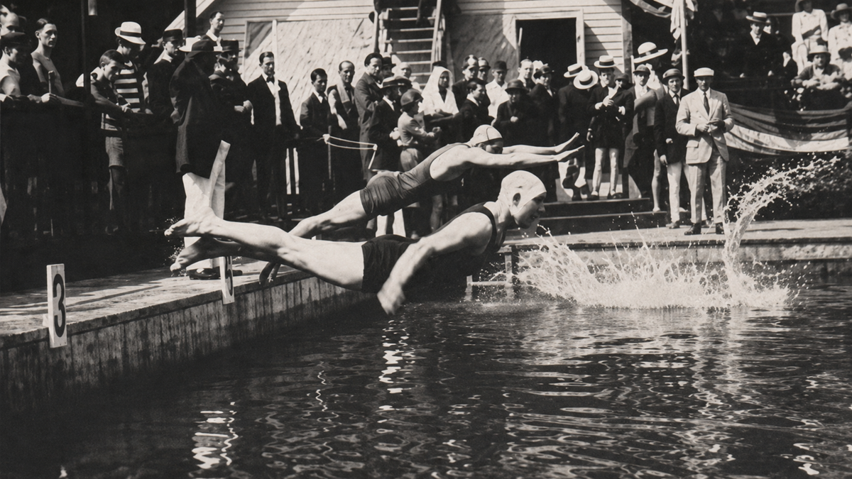 Two swimmers dive into a pool at the starting signal during a vintage swim race, while spectators in early 20th-century attire watch from the deck in the background.