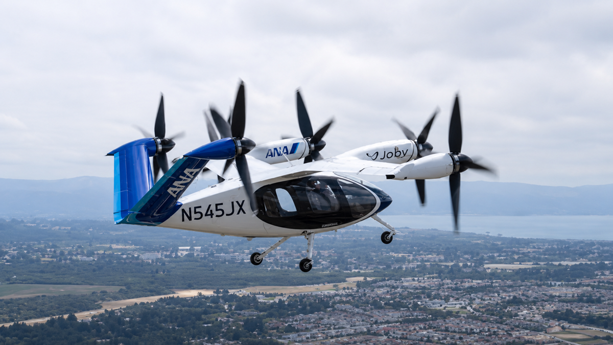 A white and blue electric vertical takeoff and landing aircraft, marked “N545JX” and labeled “ANA” and “Joby,” flies above a cityscape with hills and water visible in the background under a cloudy sky.
