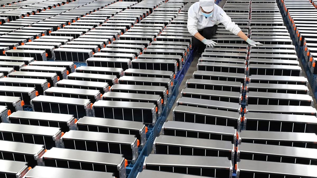 A worker in a lab coat and mask inspects large rows of rectangular battery cells, possibly for electric vehicles, neatly arranged in an industrial facility. Tech.