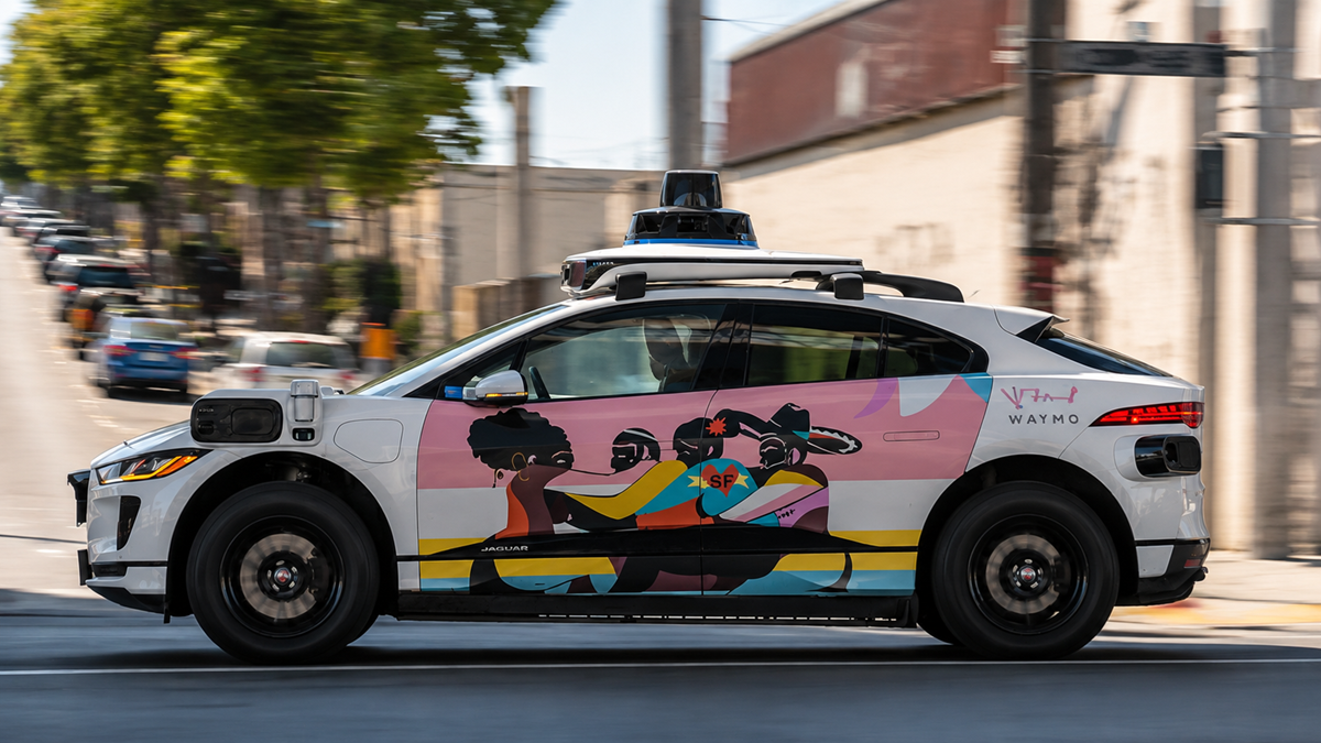 A self-driving Waymo car with vibrant, colorful artwork featuring people on its side drives on a city street during the day.